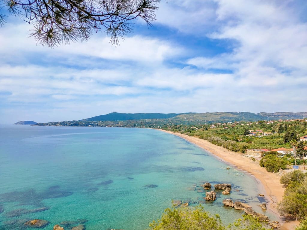 A tranquil coastal landscape featuring a sandy beach, turquoise water, and rocky outcrops. Green hills and a small town are visible in the background, beneath a partly cloudy sky. Zaga beach in Koroni