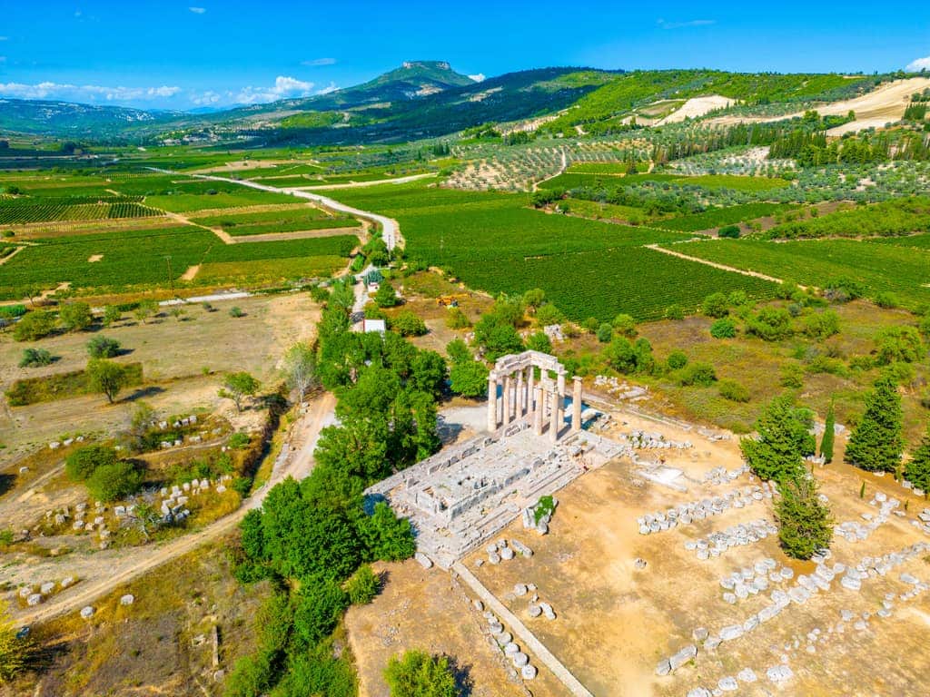 Aerial view of the ancient Temple of Zeus ruins in Nemea, Greece, featuring several standing columns and scattered stones. The site is surrounded by lush green fields, vineyards, and distant mountains, blending natural beauty with historical architecture. Vineyards and ancient ruins in Nemea, Greece