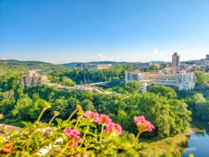 ALT text: Scenic city view framed by pink flowers in the foreground, with lush greenery and hills surrounding a mix of modern and historical buildings; notable features include a large building complex on the right and a central monument with statues, all under a bright blue sky on a sunny day; A view of Veliko Tarnovo with one of the best hotels in Veliko Tarnovo: Grand Hotel