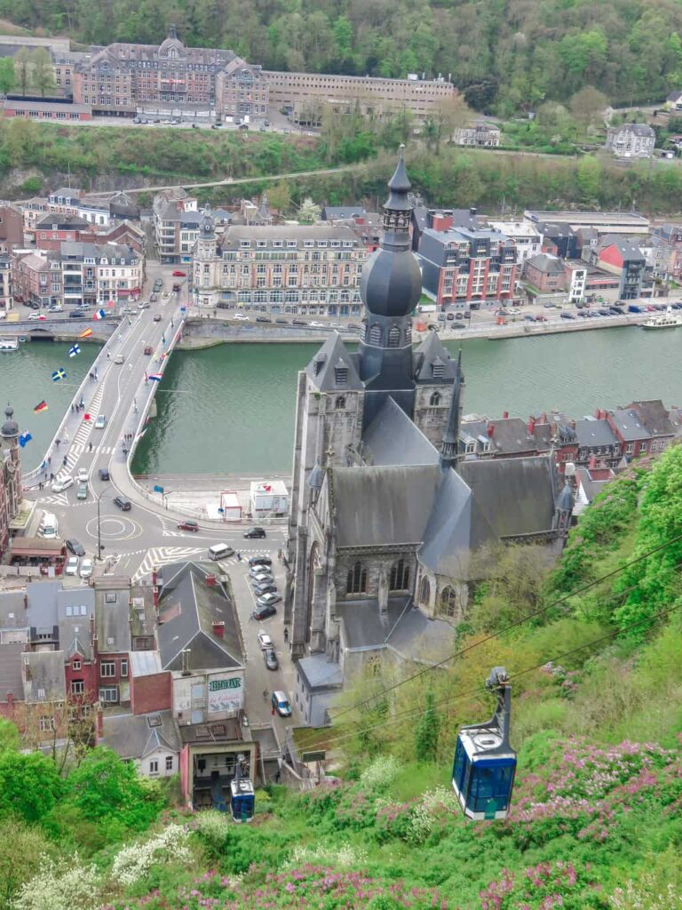 A large stone church with a tall central tower stands beside a river in a city with bridges, roads, and buildings. A cable car descends a green hillside in the foreground. View from the Citadel in Dinant