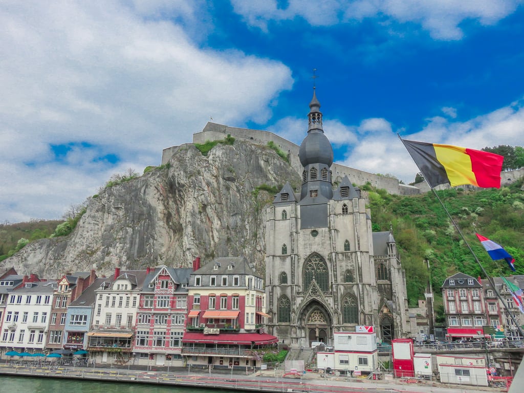 A riverside view of the Church of Notre-Dame de Dinant in Belgium, with colorful buildings, a rocky cliff, the Dinant Citadel above, and a Belgian flag in the foreground under a partly cloudy sky.