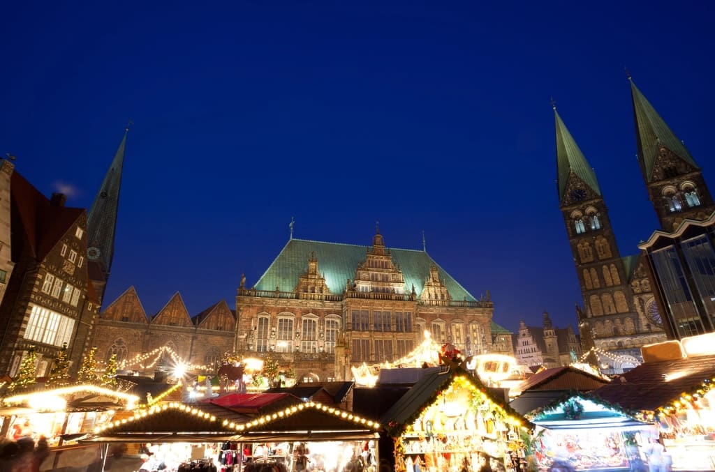 A festive market with colorful lights and decorated stalls takes place in front of a large historic building with a green roof and pointed towers under a deep blue evening sky. Bremen Christmas Market