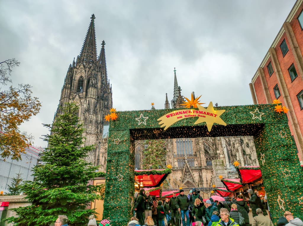 A gothic cathedral at the background with an a Christmas tree and decorated entrance to a Christmas market with text in German on a falling star: Christmas Market at the Cologne Cathedral