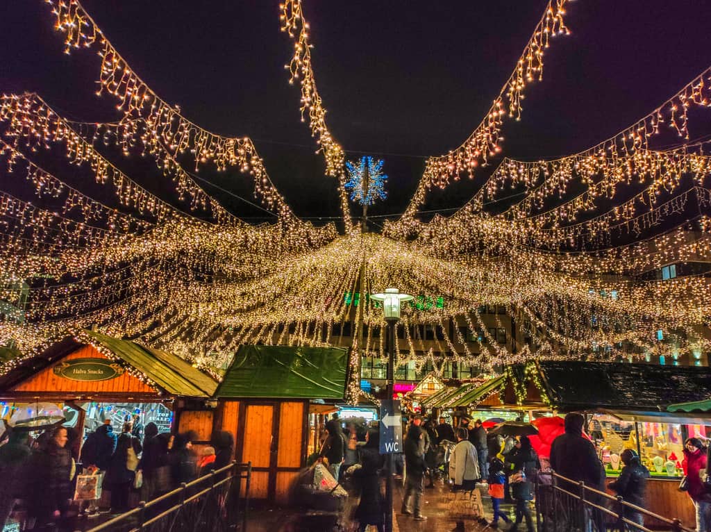 A festive outdoor market at night is decorated with numerous strings of warm white lights. Wooden stalls are set up, and people walk around shopping under the illuminated canopy. Essen Christmas Market in Germany