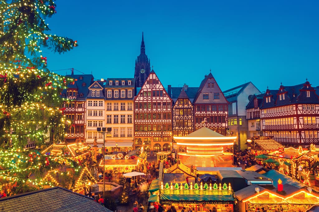 A festive Christmas market scene with colorful stalls, a large decorated tree on the left, and traditional timber-framed buildings in the background under a clear evening sky. Crowds of people fill the market area. Frankfurt Christmas Market
