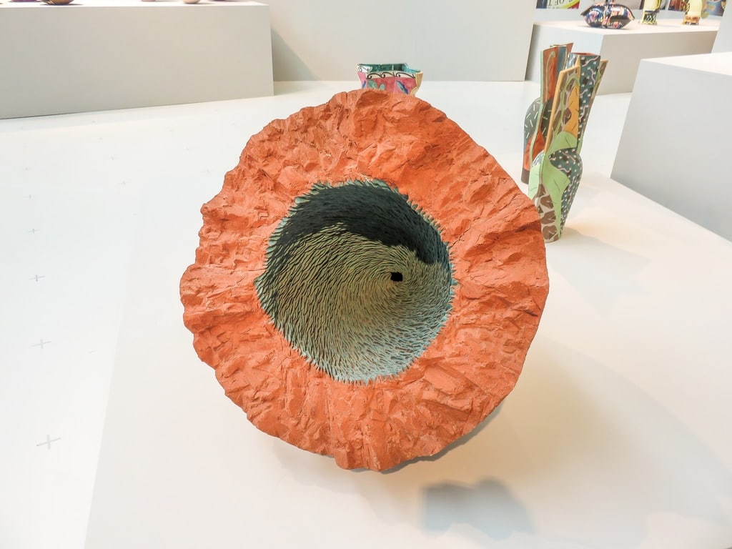 A large ceramic sculpture with a rough, orange outer surface and a textured, greenish interior is displayed on a white platform with other colorful ceramic pieces in the background. Ceramics at the Design Museum in Den Bosch