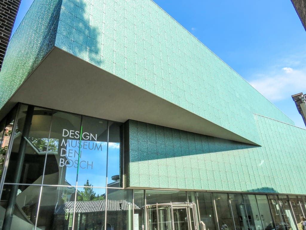 The image shows the exterior of the Design Museum Den Bosch, featuring angular modern architecture with a green-tiled facade and large glass windows, under a clear blue sky.