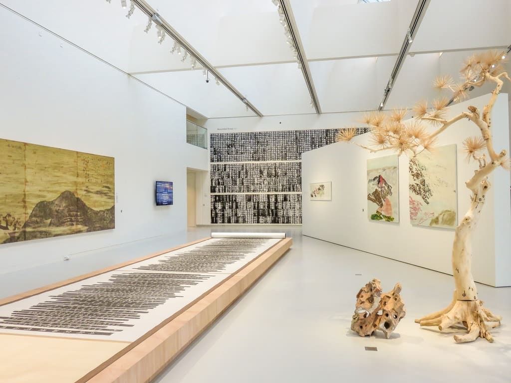 An exhibition hall with large abstract paintings on white walls, a long installation with text on the floor, a digital screen, and sculptural pieces resembling dried trees and rocks under a bright skylight. A temporary exhibition at Het Noordbrabants Museum
