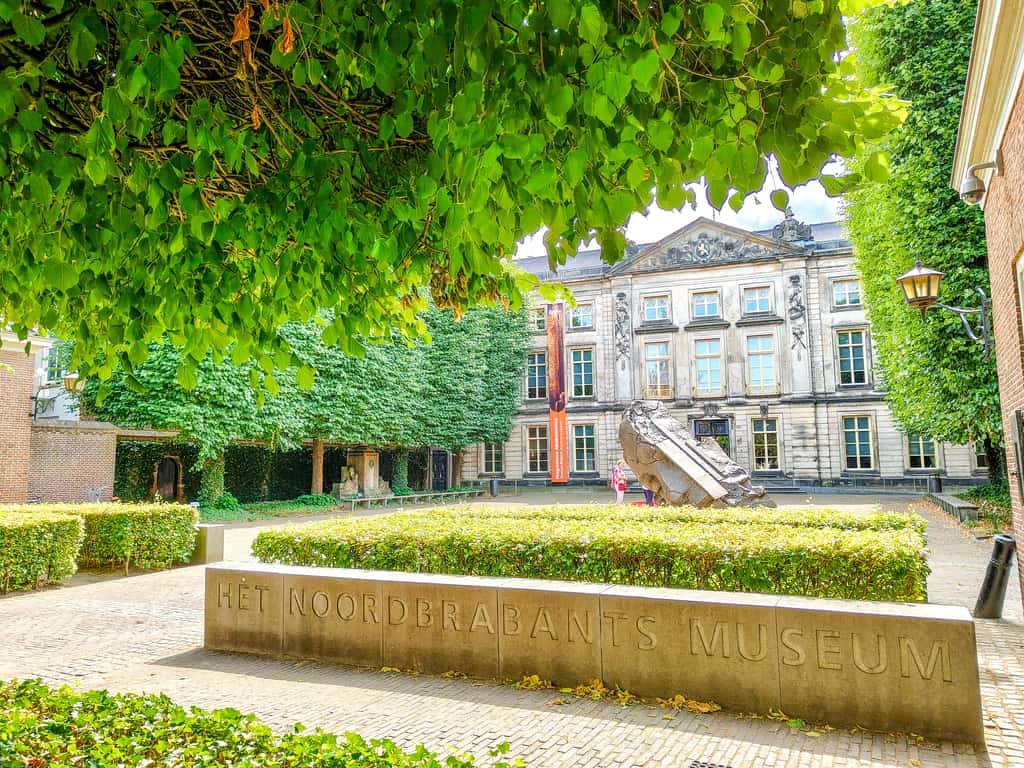 Courtyard of Het Noordbrabants Museum with a large stone sign in front, manicured hedges, leafy trees, and the museum’s historic stone building in the background on a bright day.