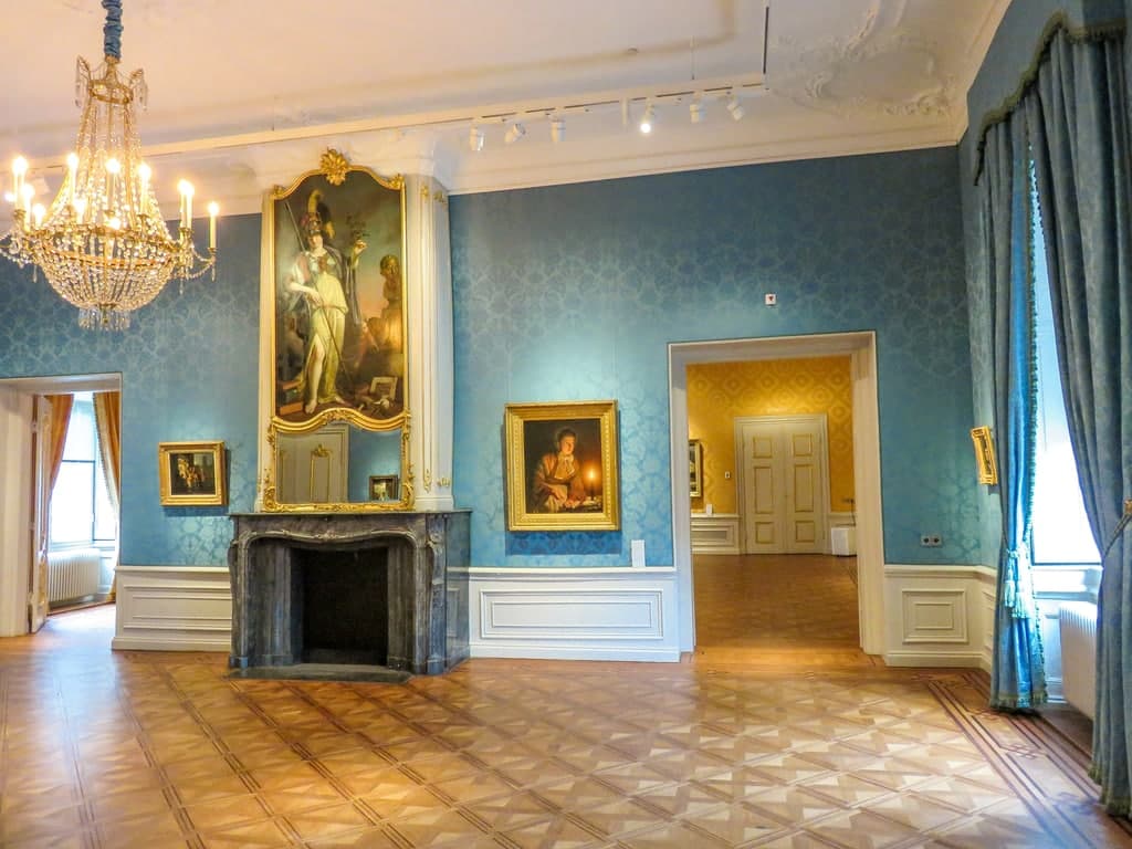 A grand room features blue wallpaper, a marble fireplace beneath a large portrait, a chandelier, parquet flooring, and several framed paintings. Tall windows with blue drapes and doorways to adjoining rooms are visible. Het Noordbrabants Museum, Den Bosch