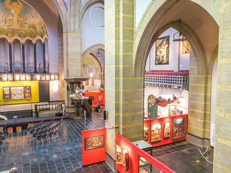 A spacious museum interior with high arched ceilings, stone columns, and display cases containing various artworks and artifacts. Rows of chairs are arranged for visitors, and large paintings hang on the walls. Hyeronimus Bosch Centre in Den Bosch
