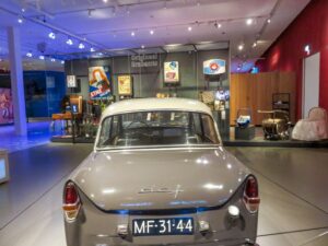 A gray vintage car with the license plate "MF-31-44" is displayed indoors at a museum, with retro signs, exhibits, and objects visible in the background.