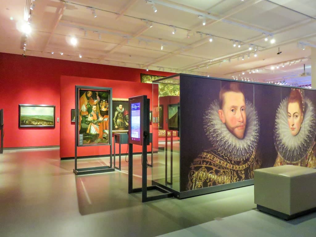 A brightly lit art gallery with red walls displays large portraits of people in Elizabethan attire, glass cases, paintings, and benches. The Story of Brabant at Het Noordbrabants Museum