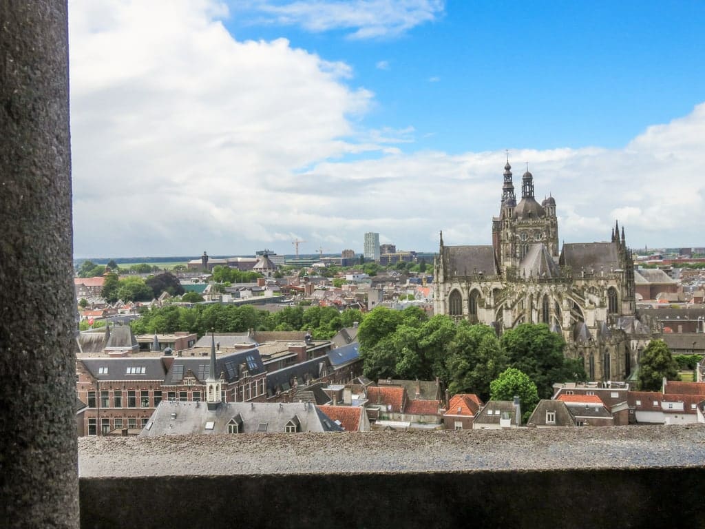 View from a stone balcony overlooking a city with red-roofed buildings, green trees, and a large Gothic-style church under a partly cloudy blue sky. View from the Jheronimus Bosch Center in Den Bosch