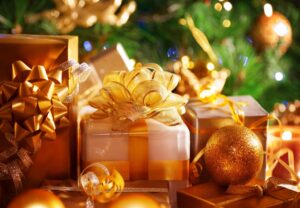 A group of wrapped gifts with gold ribbons and bows sits in front of a decorated Christmas tree with lights and gold ornaments. Best gifts for travelers