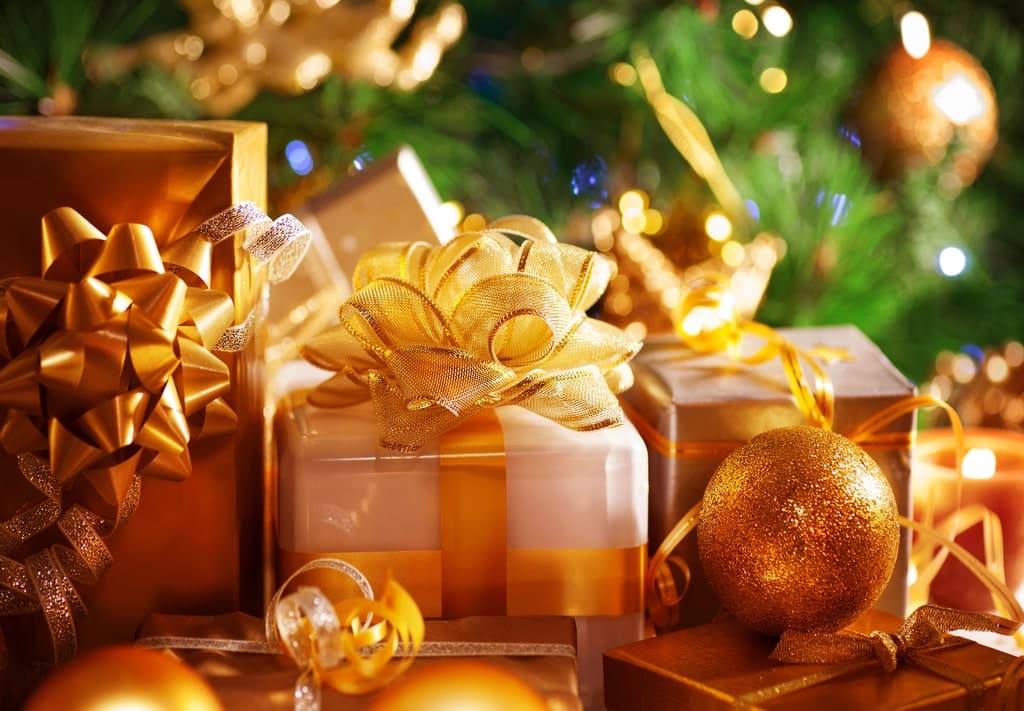 A group of wrapped gifts with gold ribbons and bows sits in front of a decorated Christmas tree with lights and gold ornaments. Best gifts for travelers