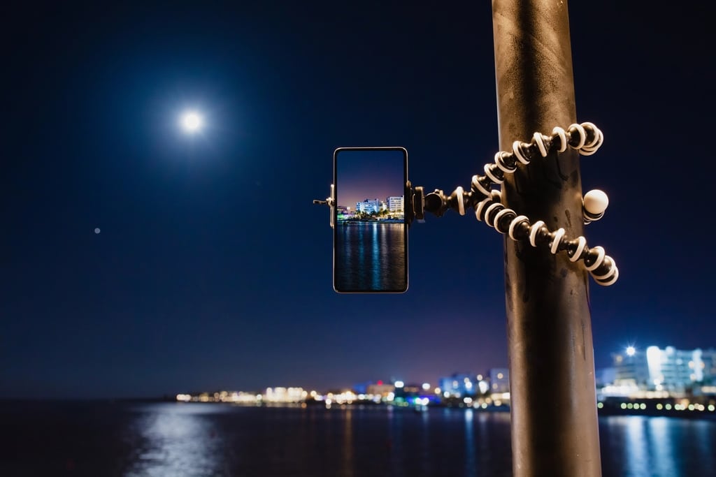 A smartphone mounted on a flexible tripod is attached to a pole, capturing a cityscape at night with buildings and lights reflected in the water; the moon is visible in the sky.