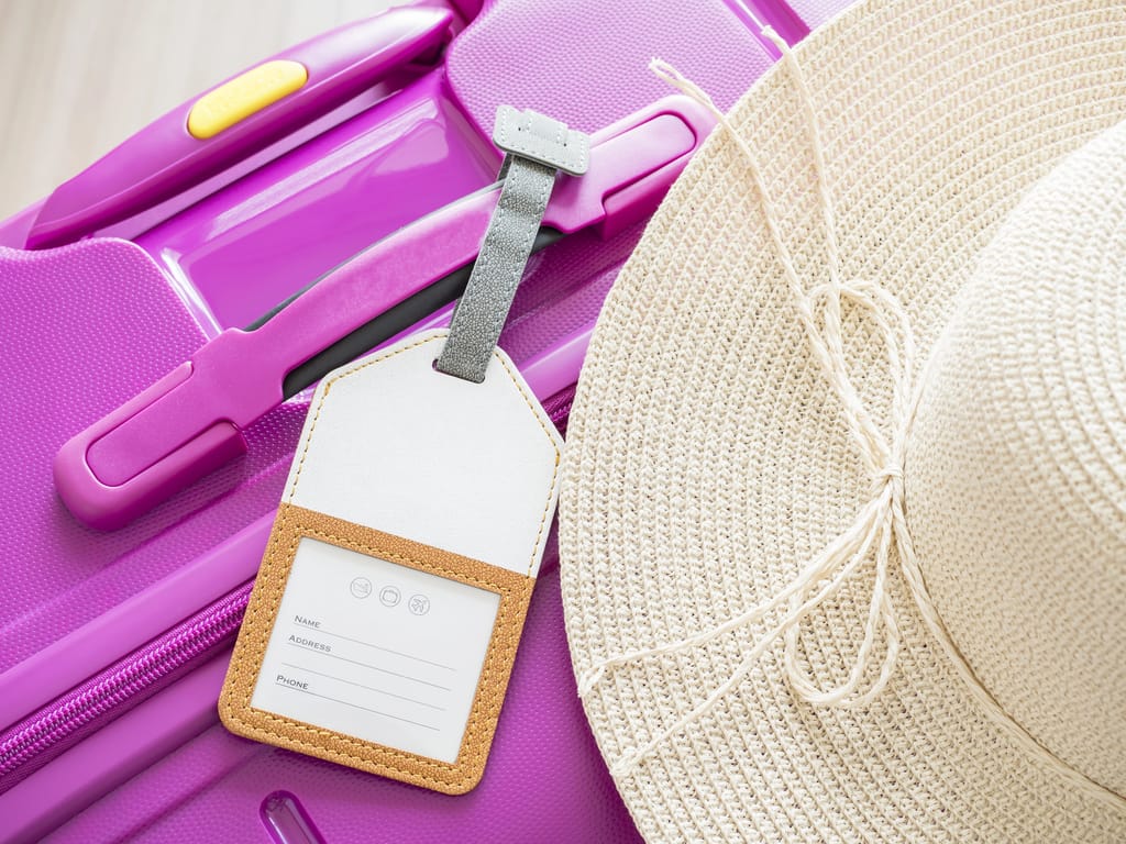 A close-up of a purple suitcase with a blank luggage tag attached and a beige wide-brimmed hat resting on top. The suitcase has a visible zipper and handle.