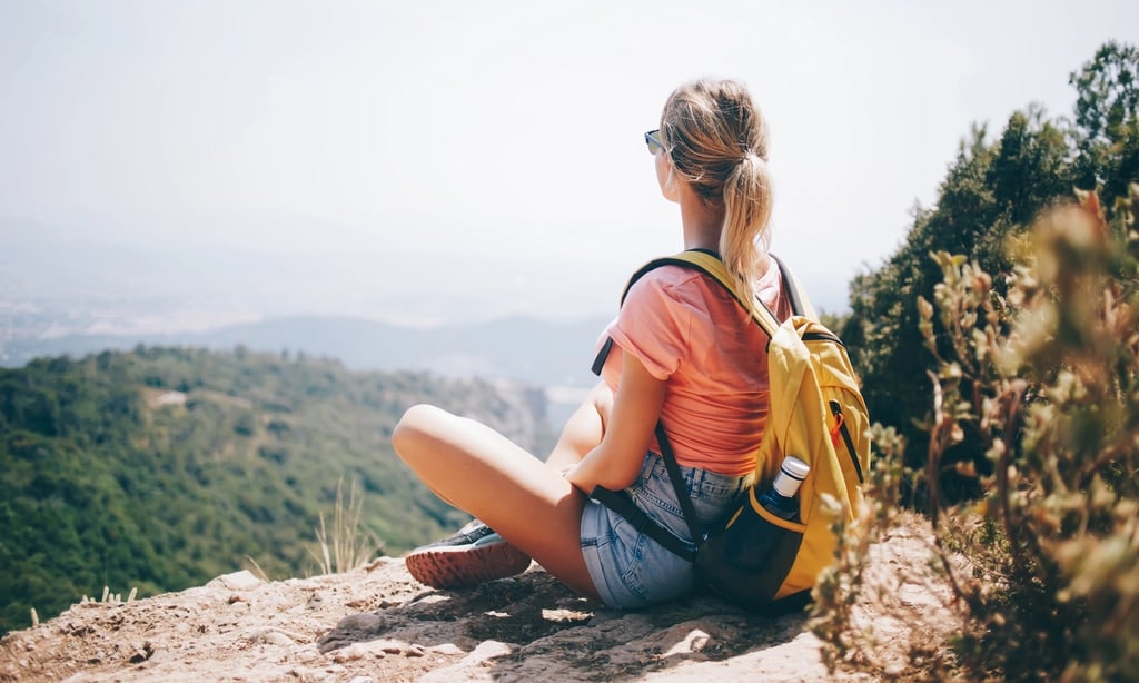 A person with a yellow backpack sits on a rocky ledge overlooking a green, mountainous landscape. They are wearing sunglasses, a pink shirt, denim shorts, and hiking boots. The sky appears clear.