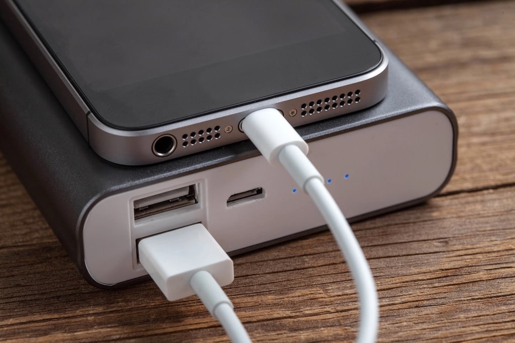 A smartphone is connected to a portable power bank with a charging cable. The power bank has USB and micro-USB ports, with indicator lights on, and is placed on a wooden surface.