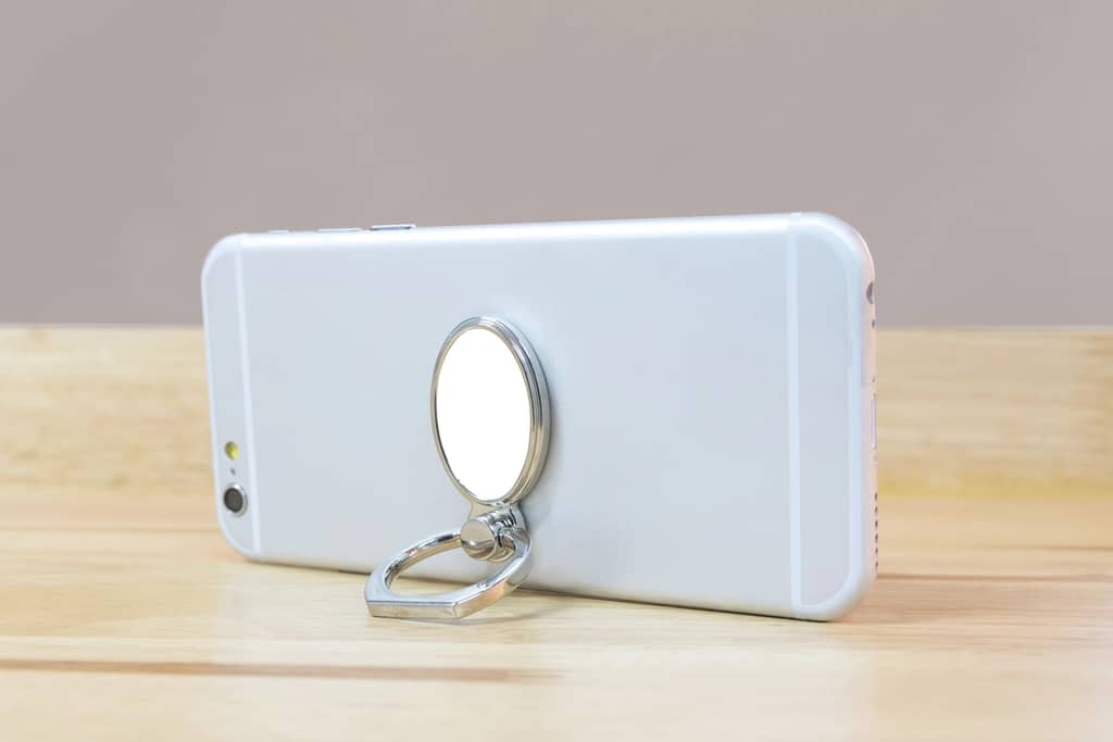 A silver smartphone with a metallic ring holder attached to the back is propped up on a wooden surface, using the ring to stand upright in a landscape orientation.