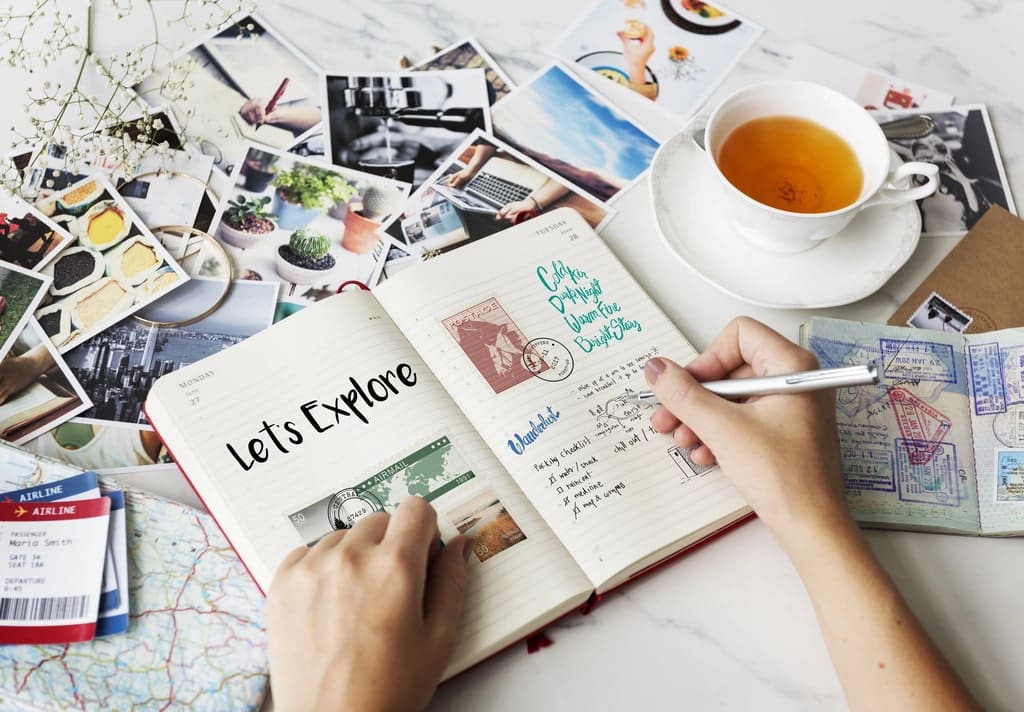 A person writes in a travel journal at a table with a cup of tea, travel tickets, postage stamps, a map, and scattered travel photographs. The journal’s open page reads "Let's Explore.