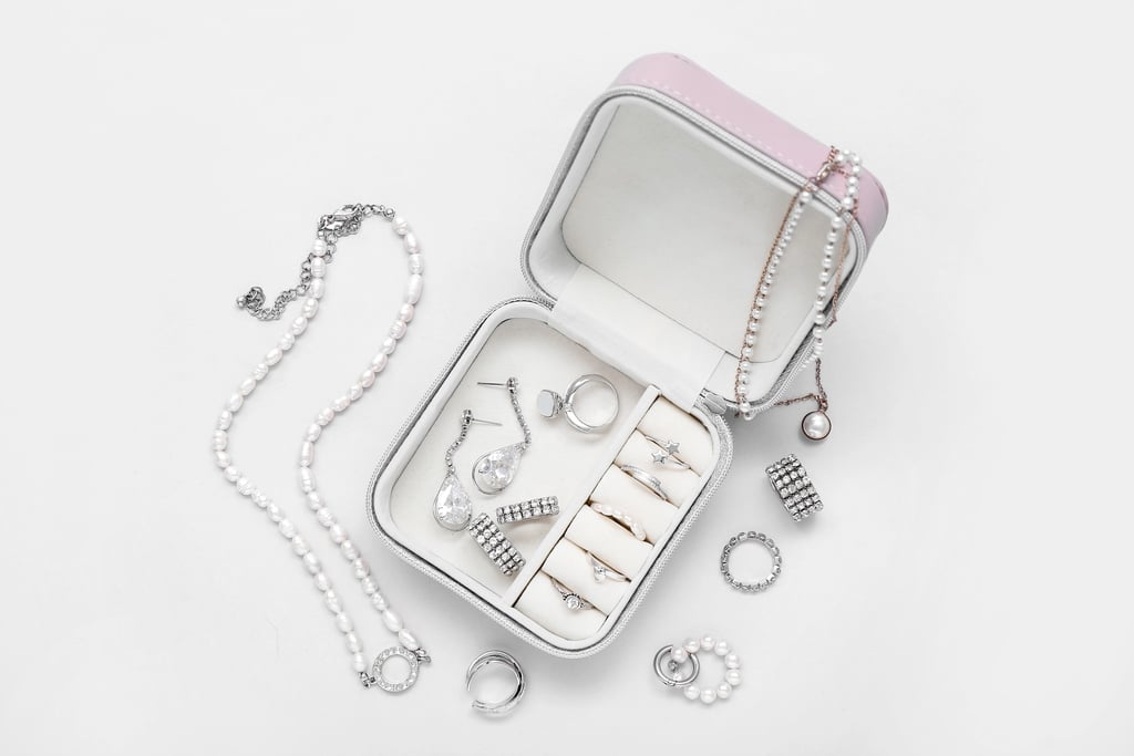 A white jewelry box with rings, earrings, and a pair of studded cufflinks. A pearl necklace, a silver chain with a pearl pendant, a ring, and a beaded bracelet are arranged around the box on a white surface.