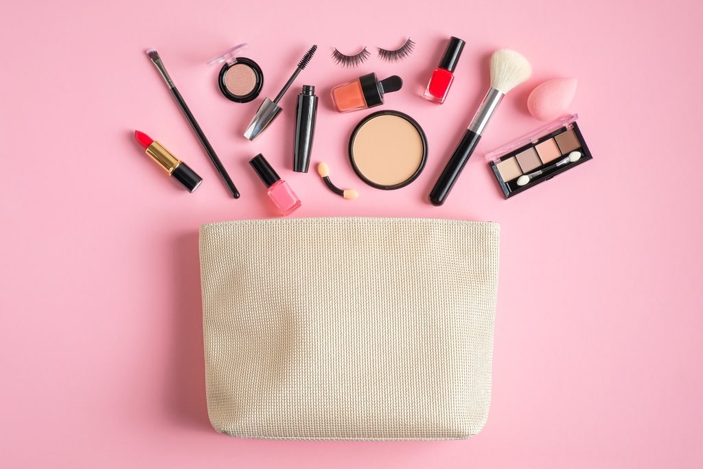 A beige makeup bag on a pink background with various makeup items arranged above it, including lipstick, nail polish, eyeshadow, brushes, mascara, false eyelashes, foundation, and a beauty sponge.