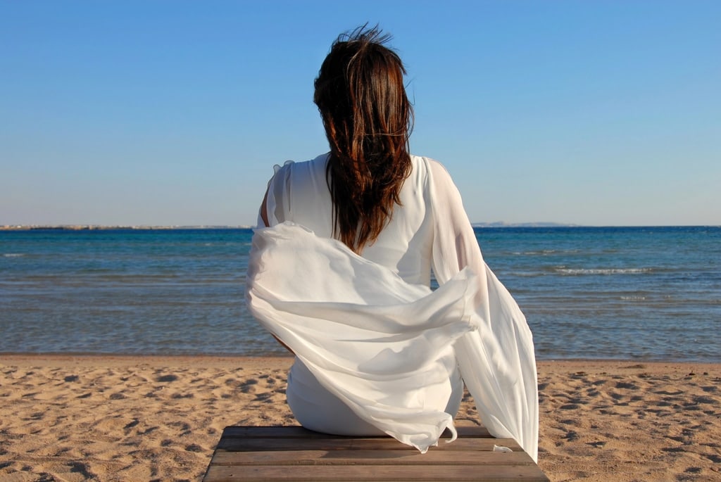 A person with long brown hair, wearing a white dress, sits on a bench facing the ocean. The sky is clear and blue, and gentle waves are visible on the sandy beach. The dress flows in the breeze.