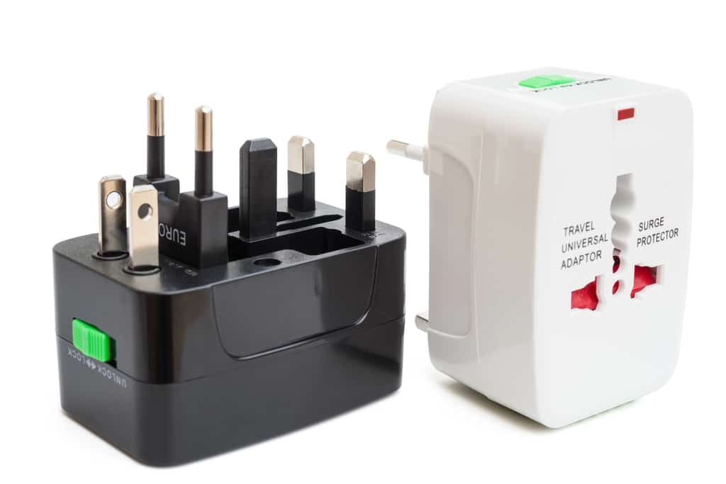 A black and a white universal travel power adapter are displayed side by side. The black adapter shows multiple plug types extended, while the white adapter has labeled sockets and a surge protector switch.