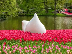 A large white abstract sculpture sits amid a field of pink tulips next to a pond, with green trees and people walking in the background. Keukenhof Gardens, the Netherlands