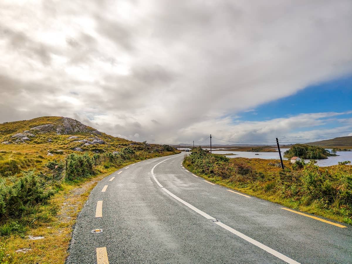 A winding road curves through a rural landscape with grassy hills on the left and a body of water on the right under a partly cloudy sky. Telegraph poles line the road, and the area appears peaceful and remote. Ireland without a car road trip