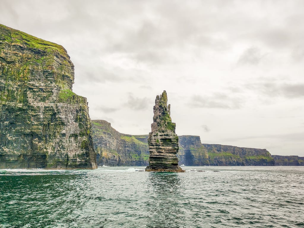 A tall, jagged sea stack rises from the sea near steep, green-topped cliffs under an overcast sky at the Cliffs of Moher in Ireland.