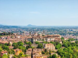 A panoramic view of Bergamo, Italy, showing historic buildings with red-tiled roofs, church domes, towers, and lush green trees, with the distant mountains and blue sky in the background.