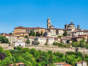 A panoramic view of Bergamo Alta in Italy, showing historic buildings with red-tiled roofs, a bell tower, and a church dome, all surrounded by lush green trees under a clear blue sky. Citta Alta bergamo