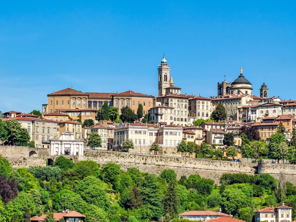 A panoramic view of Bergamo Alta in Italy, showing historic buildings with red-tiled roofs, a bell tower, and a church dome, all surrounded by lush green trees under a clear blue sky. Citta Alta bergamo